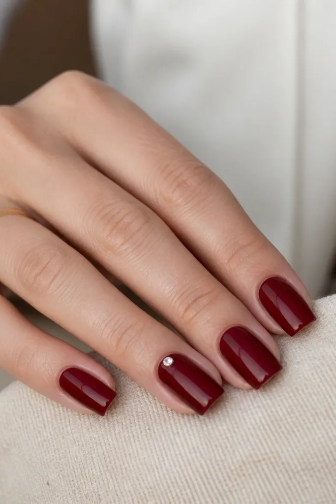 All nails solid glossy dark cherry red square; one nail has a single small clear crystal near the cuticle, held flat.