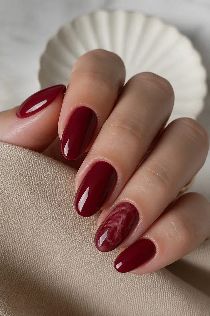 Three nails solid glossy dark cherry red; the ring finger has a dark cherry marble pattern with faint lighter maroon swirls, glossy.