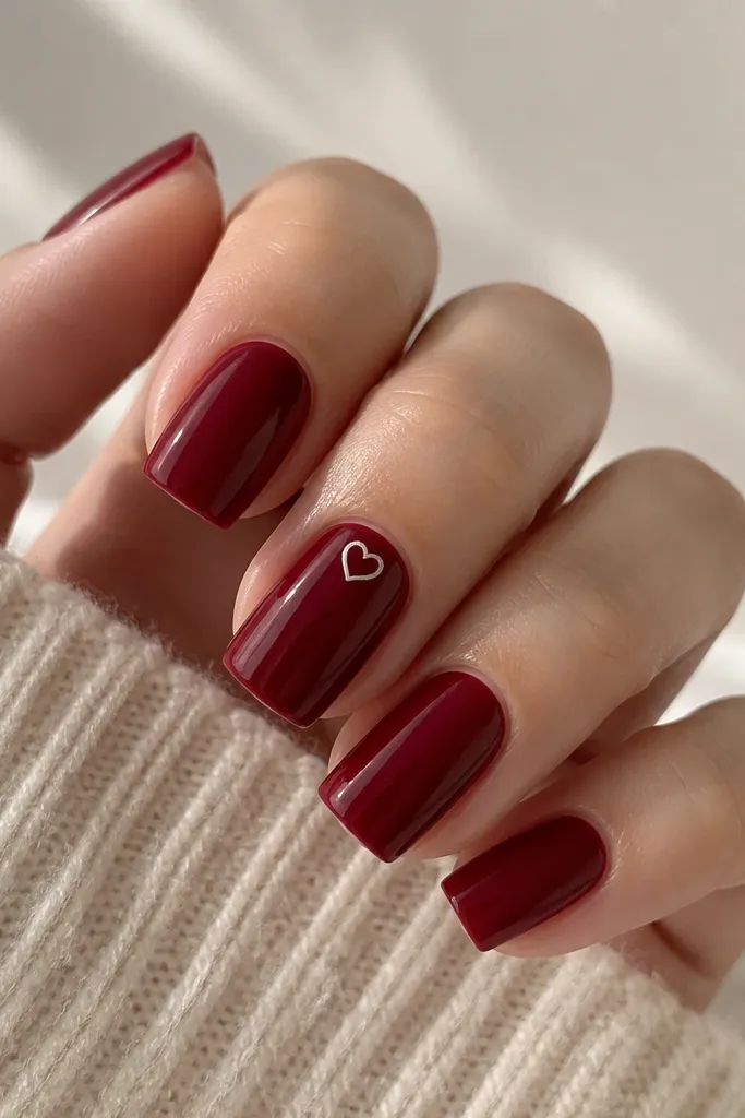 Square nails solid glossy dark cherry red; the accent nail has a small thin white heart outline near the cuticle center.
