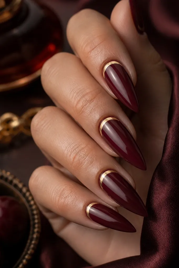 Stiletto nails in dark cherry red with a thin gold line forming a half-moon near the cuticle. The gold is crisp and reflective, with a clean gap between the line and the cherry base.