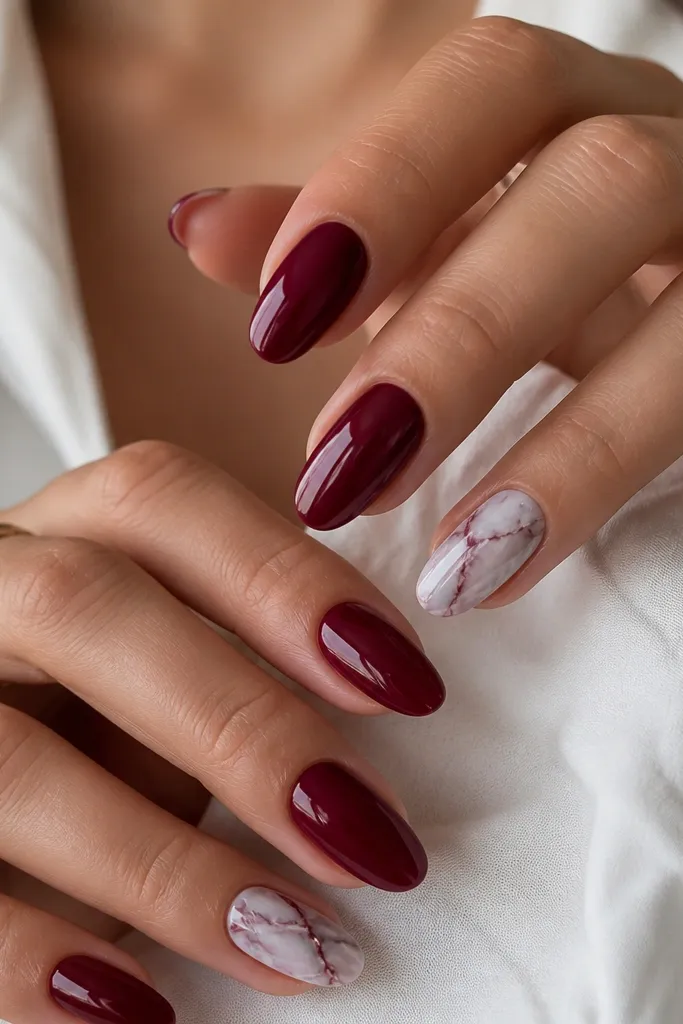 Dark cherry red nails with marble veins in one or two accent nails. The veins look like thin, feathery lines in deep burgundy and cool gray, branching from the center toward the sides. Other nails stay solid cherry red. Glossy finish.