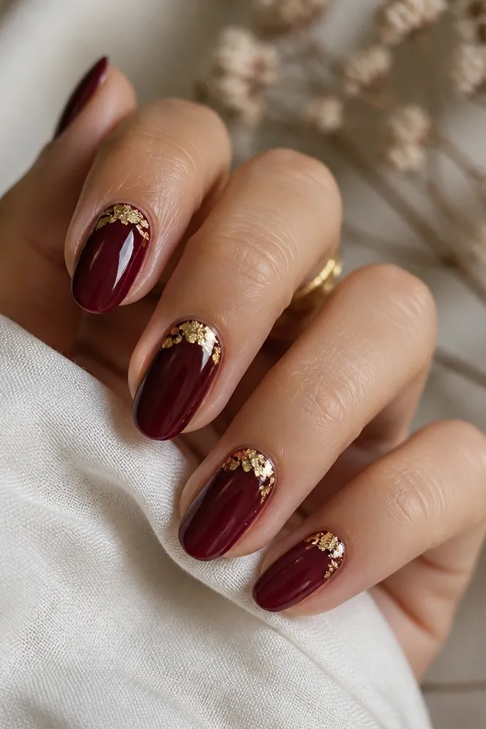 Nails in dark cherry red with a small gold leaf triangle placed in the upper corner near the cuticle on each nail. Gold leaf is irregular and slightly scattered. Rest of nail is solid cherry red with glossy shine.