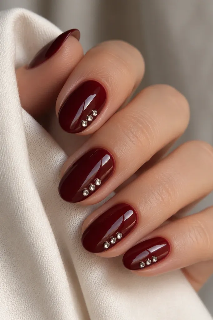 Dark cherry red nails with three small rhinestones stacked diagonally along one sidewall of each nail, starting near mid-nail. Rhinestones are clear and catch light. Glossy finish.