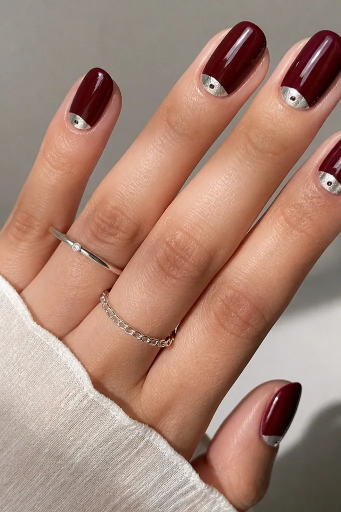 Dark cherry red nails with a reverse French half-moon at the cuticle in silver chrome. On each nail, one tiny silver dot sits in the center of the half-moon. Glossy finish.