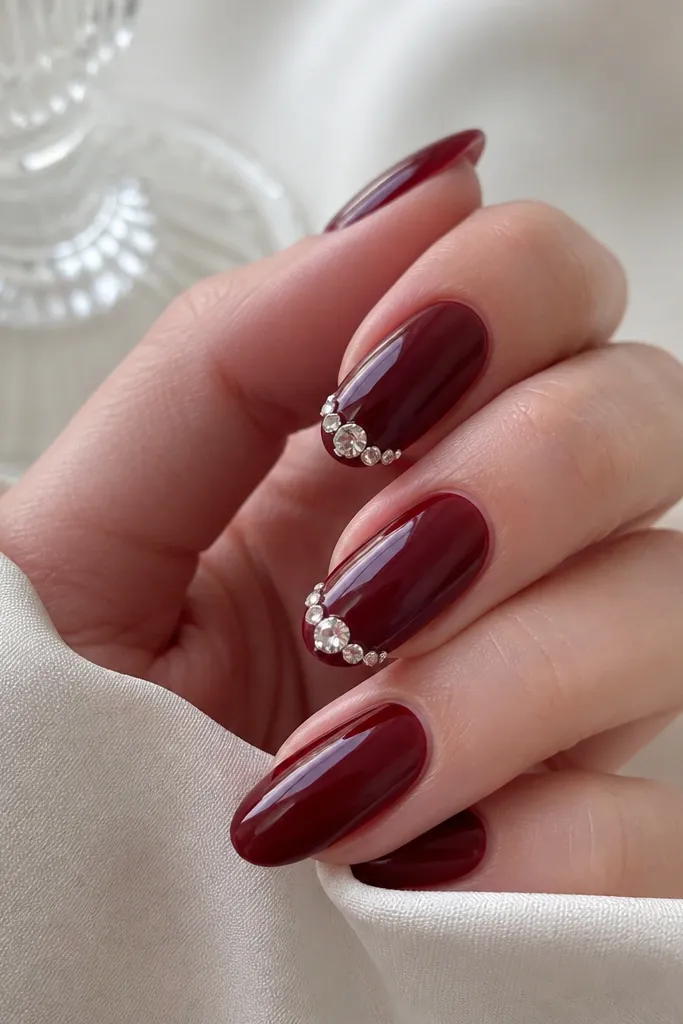 Dark cherry red nails with a cluster of clear crystals near the tip on two nails. The crystals are arranged in a small arc, with the largest crystal at the center. Other nails are solid cherry red. Glossy top coat over crystals.