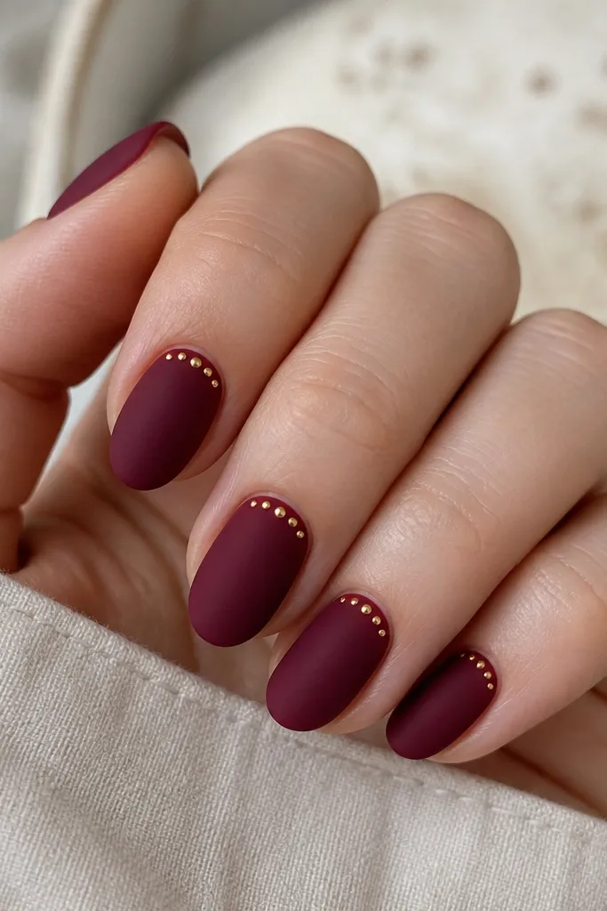 Matte deep cherry nails with tiny gold micro dots clustered near the cuticle on one side of each nail. The dots are evenly spaced and the rest stays matte; short almond shape.
