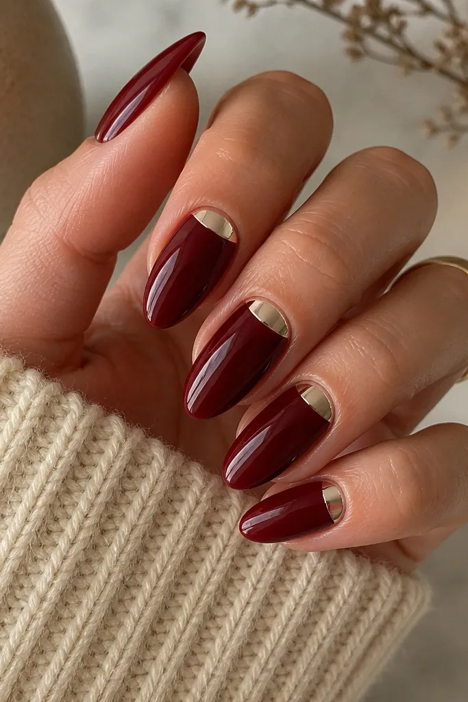 Fully glossy dark cherry nails with a half-moon chrome accent at the cuticle on each nail. The chrome is silver-champagne and sits in a neat curve; almond shape.