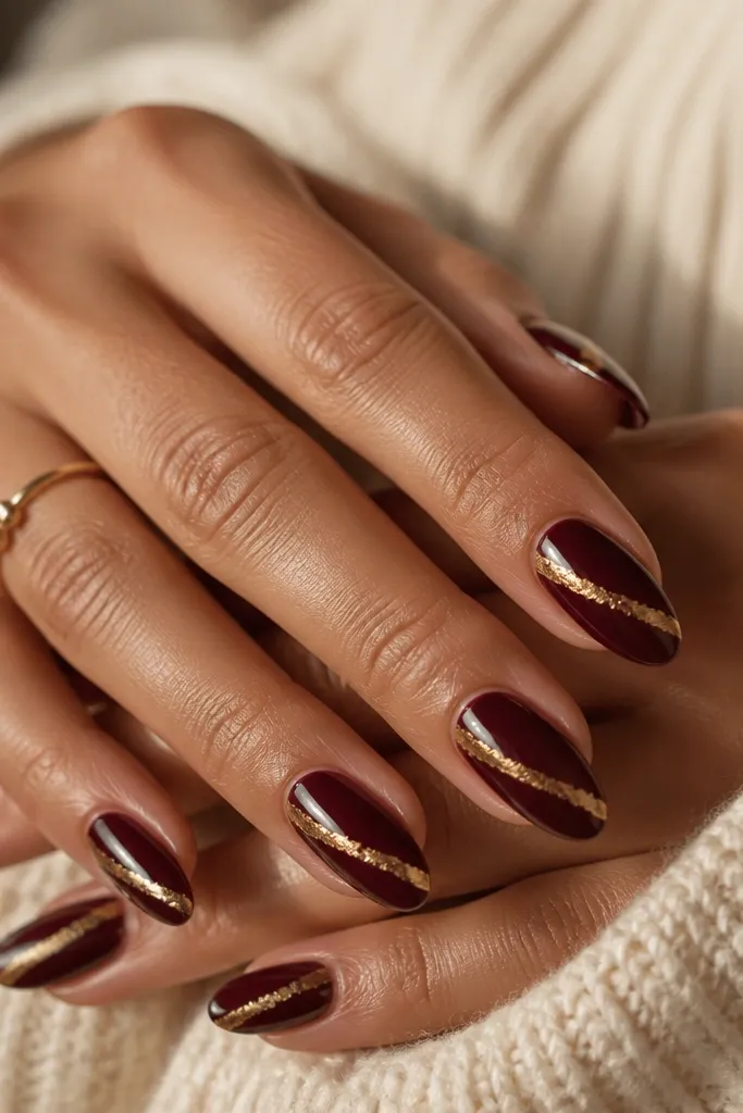 Glossy dark cherry nails with a single diagonal strip of gold foil across one side of each nail. The foil is thin and irregular at the edges; almond shape.