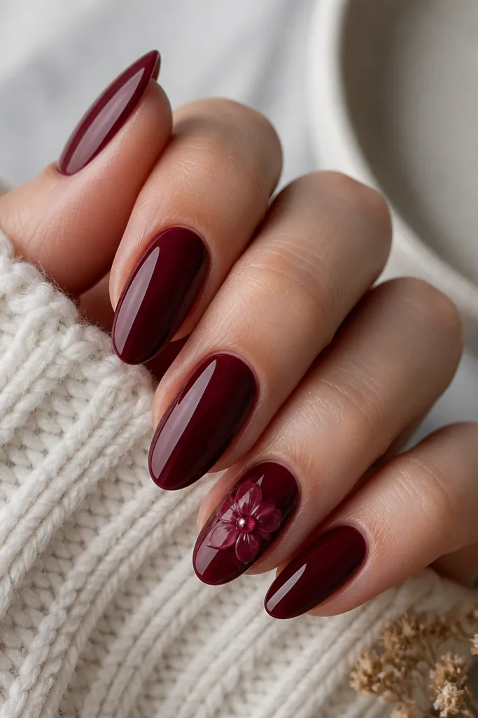 Most nails are solid glossy dark cherry. One accent nail has a small bloom flower painted in lighter cherry petals, with a shiny 3D clear gel dot in the center; almond shape.