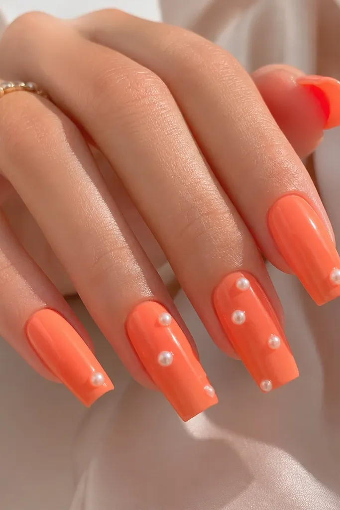 Long square nails in coral orange glossy gel. On two nails (alternating), there are small 3D pearl studs placed slightly above the center, spaced like floating dots. Other nails stay plain or have one pearl near the tip. Glossy and clean.