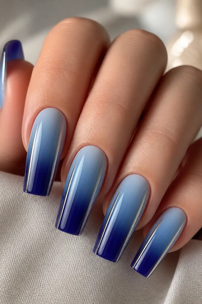Long square nails with a gradient from light blue near the cuticle to sapphire blue at the tip. On both sides of each nail, thin silver mirror lines run vertically from mid-nail to the tip. Center stays gradient and glossy.