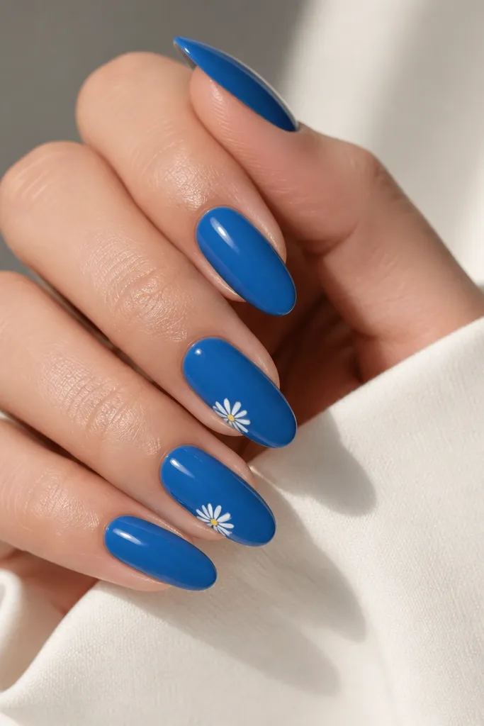 Classic blue glossy almond nails with one or two nails featuring a tiny white daisy flower near the tip. The rest are solid blue.
