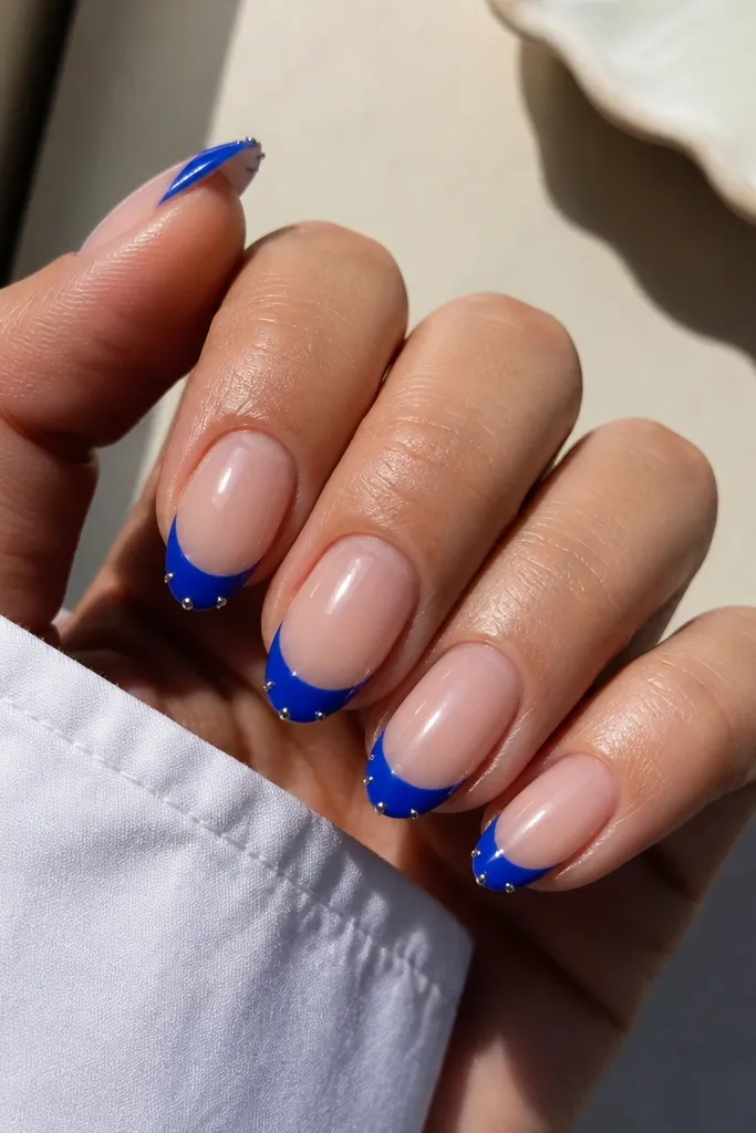 Medium almond nails in sheer nude base with bold cobalt blue French tips. At the outer corners of each blue tip, there are tiny silver studs (one each side), evenly placed. Glossy finish, clean cuticles.