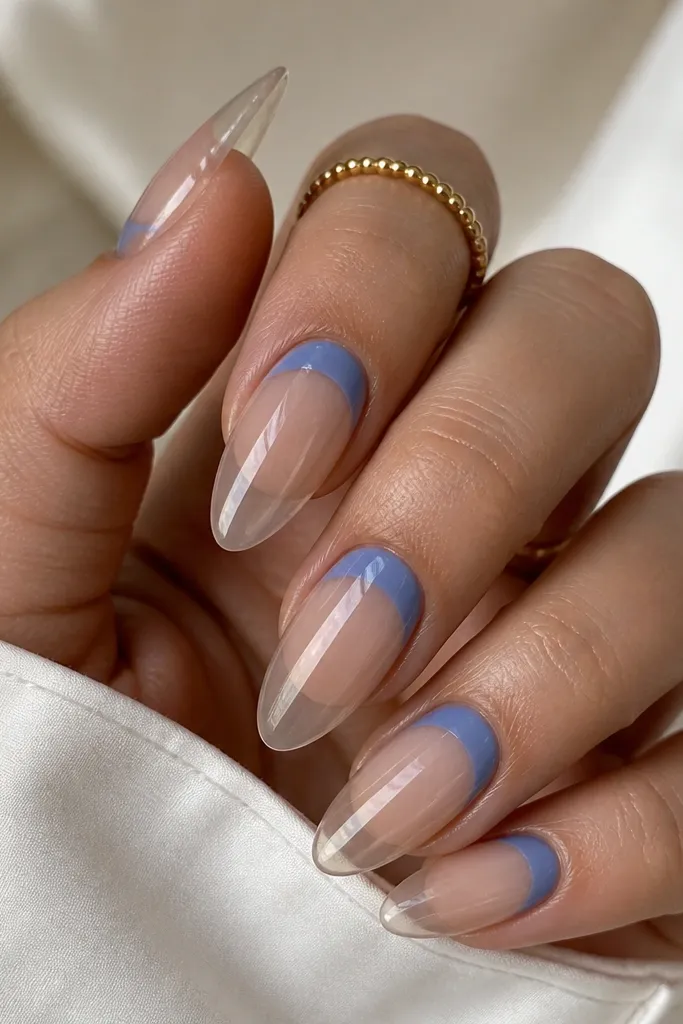 Almond nails with a clear base. The blue is painted as a reverse French along the cuticle area in periwinkle, forming a curved band near the nail bed. The free edge is clear, and the overall look is glossy.