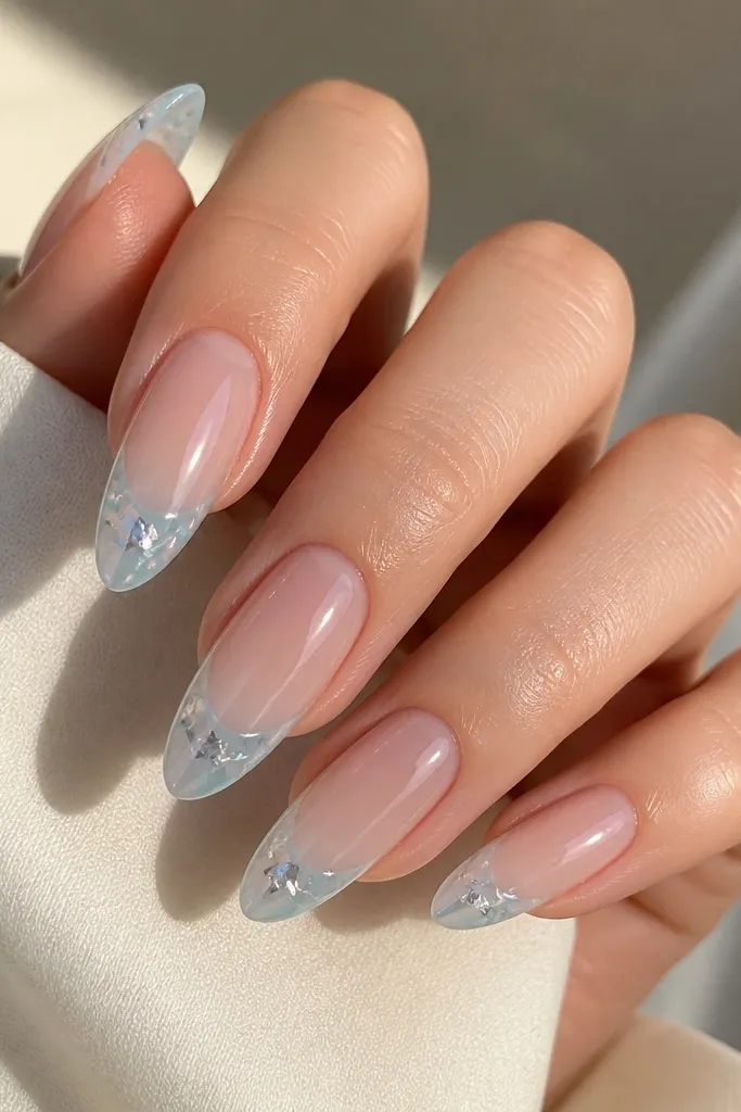 Long almond nails with a sheer nude base. French tips are icy blue with scattered silver foil flakes embedded in the blue. The flakes catch light and look like irregular shards. Glossy finish.