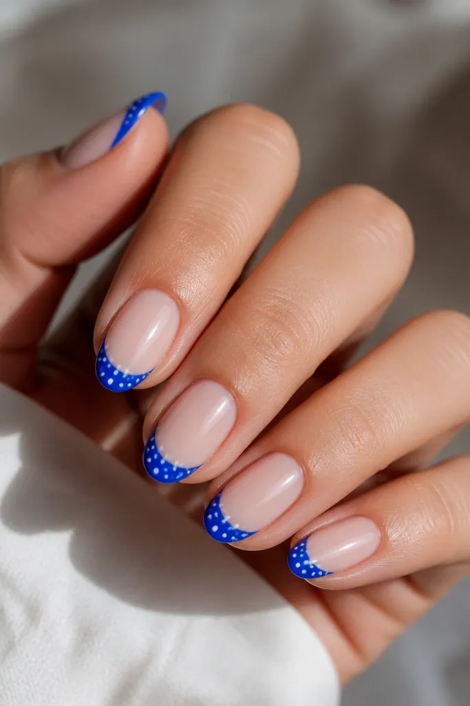 Short almond nails with a sheer nude base. Cobalt blue French tips are glossy, and there are tiny white dots scattered across the blue tip like confetti. The dots are small and evenly distributed.