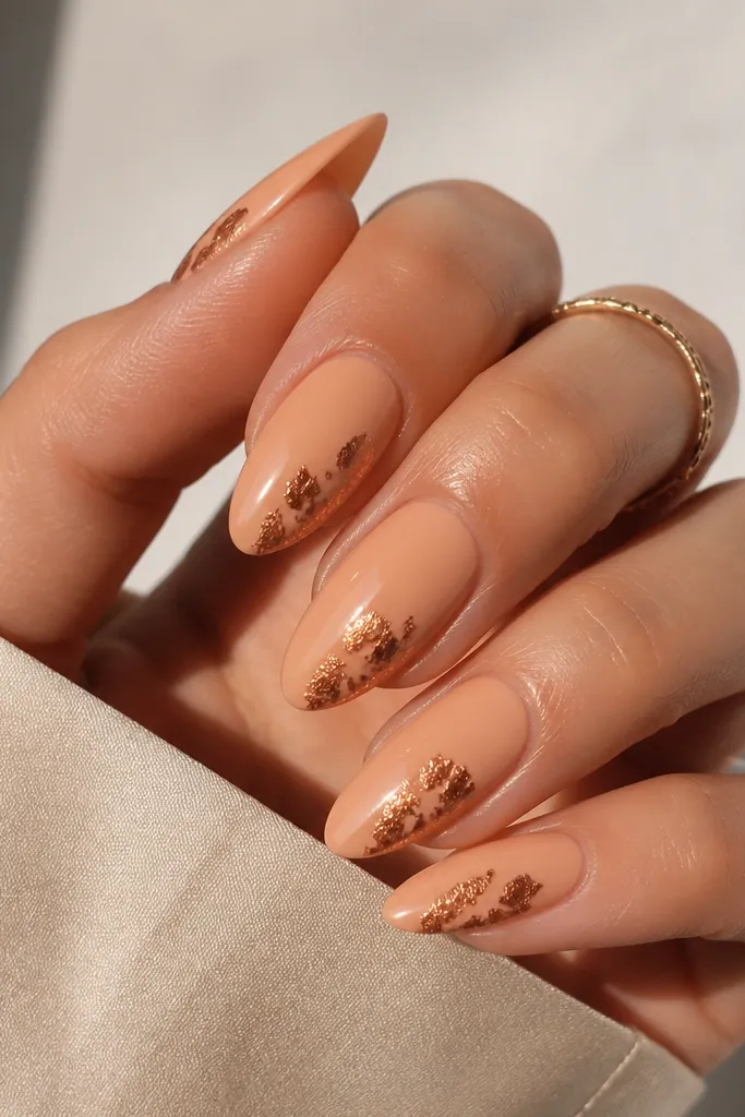 Almond nails in a creamy peach base. Bronze foil leaf flicks are placed on one side of each nail, like small torn leaf pieces, with glossy top coat over the foil.