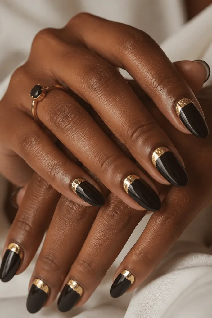 Medium almond nails with a deep espresso black base. A thin gold leaf half-moon sits right under the cuticle on each nail, with irregular leaf edges and a glossy finish over the leaf area.