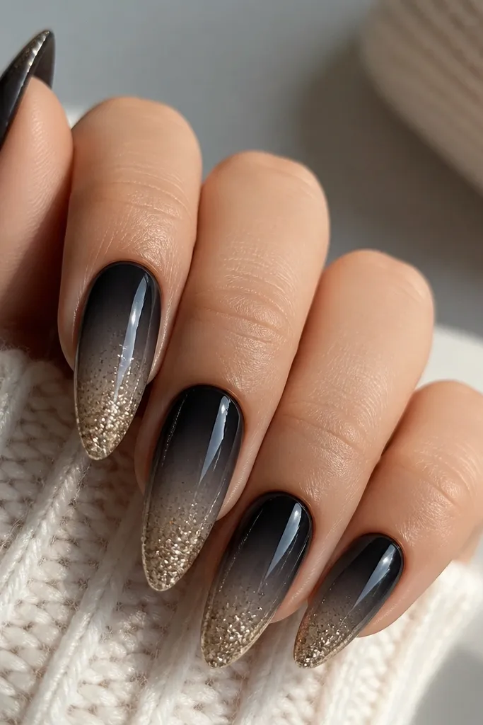 Long almond nails fading from matte black at the cuticle to a soft smoky gray mid. The tips are packed with fine gold glitter, with a smooth ombré transition and glossy top coat.