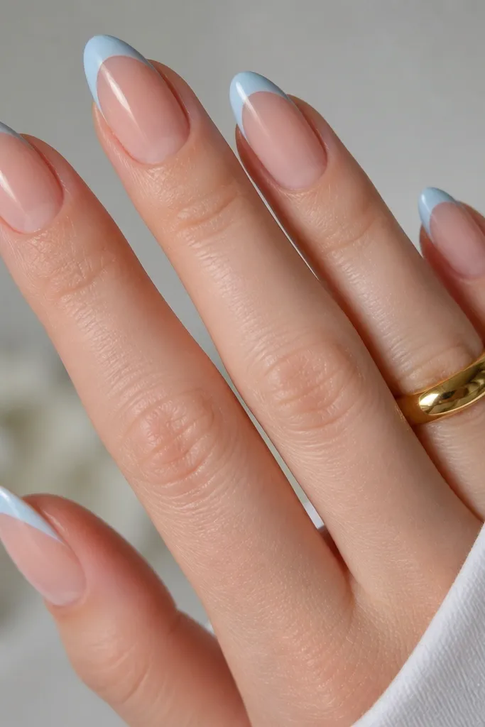 Almond nails with nude bases and light blue French tips. On the ring finger, the tip is a different light blue pattern: tiny white diagonal stripes over the blue, glossy.