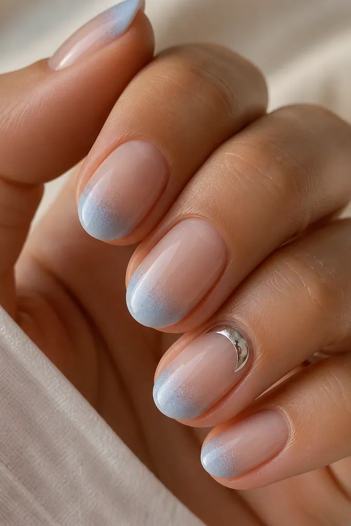 Close-up of short almond nails with a clear, glassy nude base. The French tips are coated in misty powder-blue that fades slightly at the smile line, then gets a frosted, icy matte finish only on the tip area. One nail has a tiny silver chrome crescent near the cuticle.