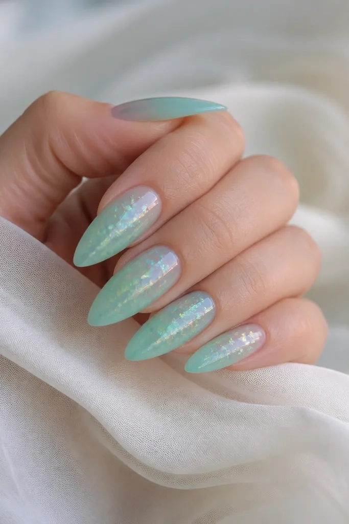 Long almond nails painted in a translucent seafoam jelly, with scattered iridescent holo flakes concentrated near the center and fading toward the cuticle; glossy top coat.