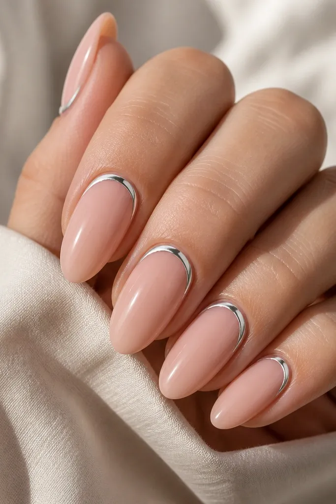 Almond nails with a nude base. Instead of a tip, there's a thin silver chrome outline following the cuticle curve, like a reverse French. The outline is mirror-bright and evenly spaced from the skin.