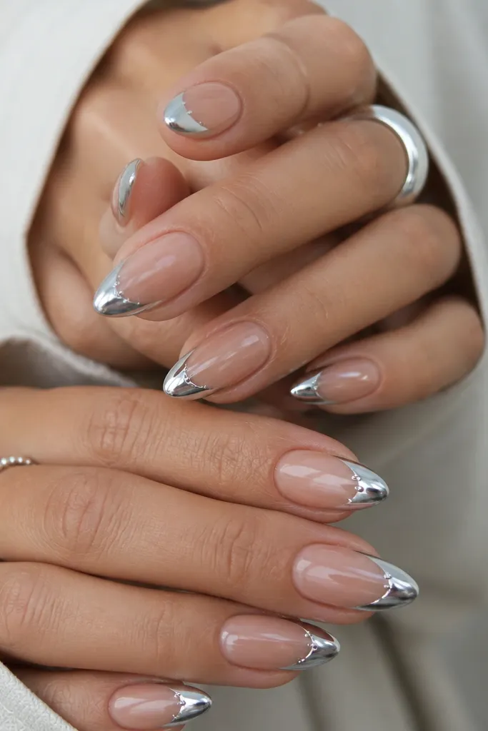 Almond nails with a nude base and mirror silver chrome French tips. Along the French smile line, there are tiny clear micro studs placed in a short row - not a full line, just a few evenly spaced points. Everything is glossy and clean.