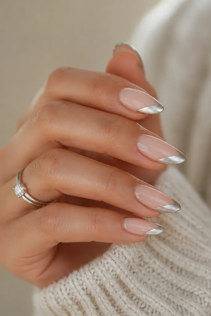 Almond nails with a sheer nude base. The French tip is silver chrome but angled diagonally from one side to the other, creating a modern slant. The chrome line is crisp and mirror-bright.