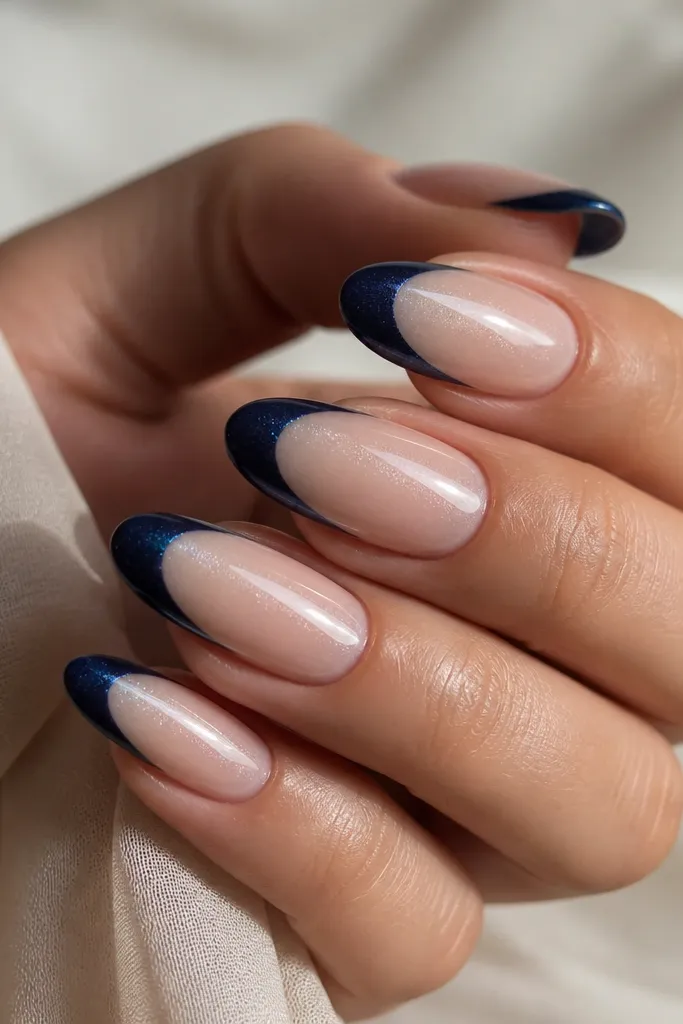 Long oval nails with a pearlized nude base that has a soft shimmer. The French tips are navy with a pearl finish, not fully metallic, and the smile line is sharp. Light reflects in tiny points across the navy.