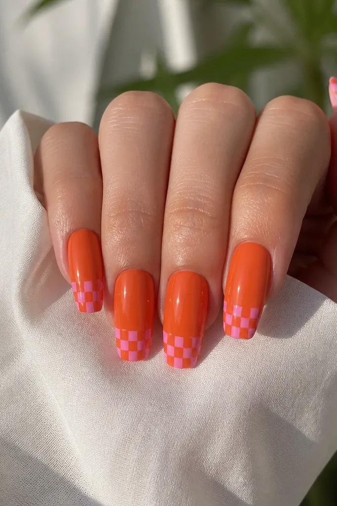 Medium coffin nails. Orange base with a checker-like tip made from alternating small pink and orange squares. The squares sit in a tight grid along the free edge. Glossy finish.