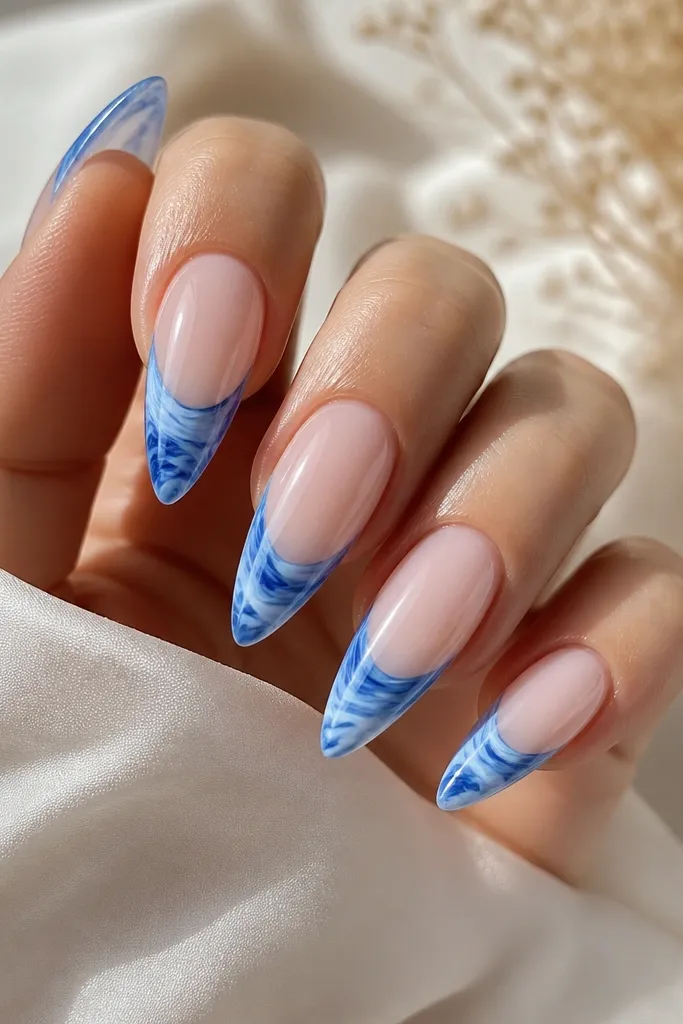 Long almond nails with icy pink translucent base. Blue French tips look marbled with white and darker blue veining, glossy and swirled.