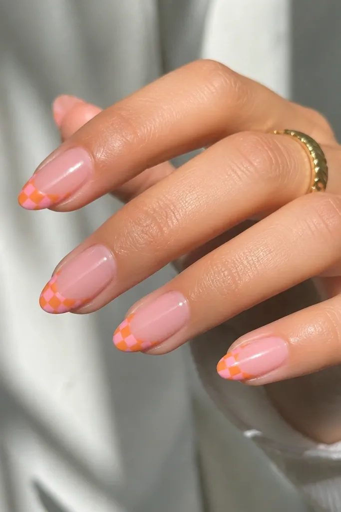 Medium almond nails with milky pink base. On the tips, a tiny checker pattern alternates orange and pink squares in a small band. Glossy finish.