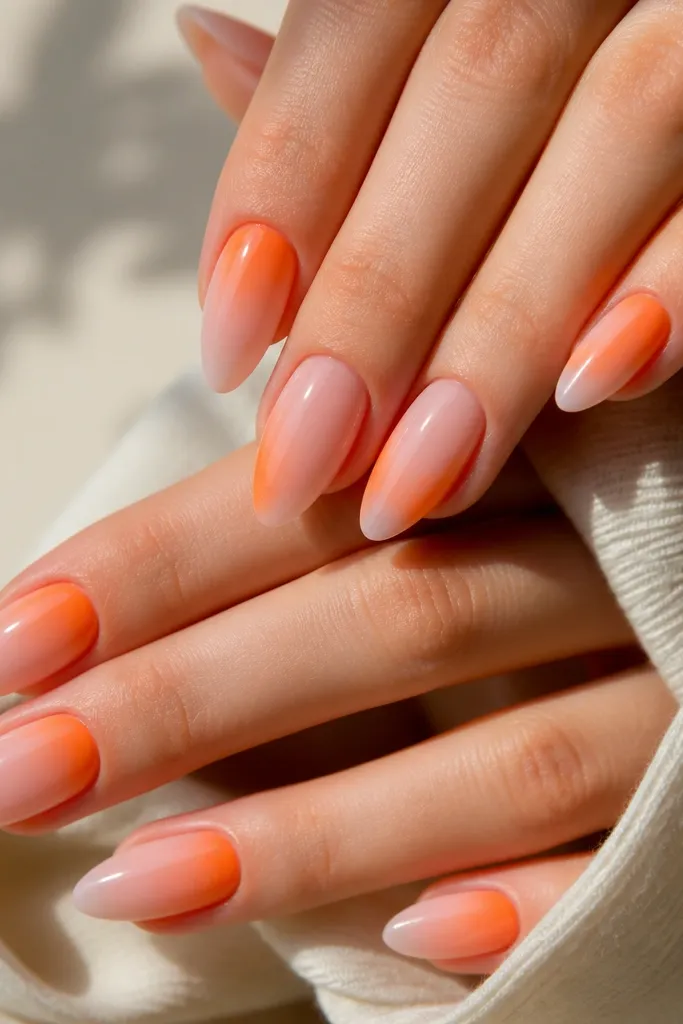 Almond nails with a sheer blush pink base fading into a bright tangerine orange glow at the lower cuticle and outer corners; glossy finish with clean, rounded cuticle edges.