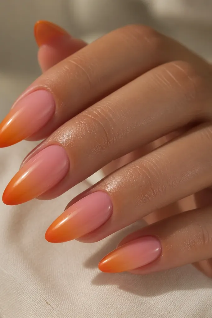 Long almond nails. The aura is peony pink near the cuticle, fading into a soft warm orange. The very tips have a deeper burnt orange ember tone, still blended rather than a hard line. Glossy and smooth.