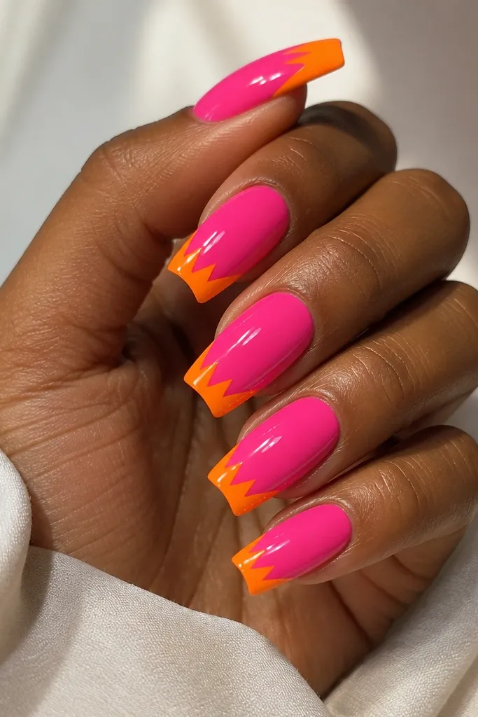 Coffin nails painted hot pink. The French tips are orange but shaped like a zig-zag across the tips, with clean lines and alternating angles. The top coat is glossy and the zig-zag sits evenly on each nail.