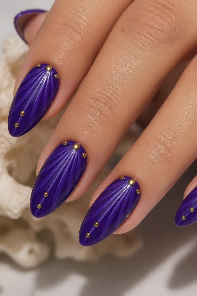 Medium almond nails. Base is royal purple. Shell ridges are slightly lighter royal tones. A few small gold pearl dots sit along the ridge lines near the cuticle and mid-nail. Glossy topcoat.