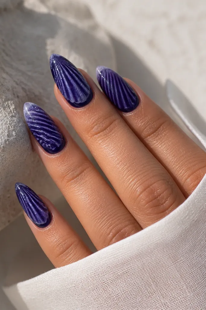 Long almond nails. Base is deep indigo. Shell ridges are drawn in darker indigo and lighter purple. A fine silver shimmer dust sits lightly over the ridges, plus a faint milky white foam edge at the outer ridge line.