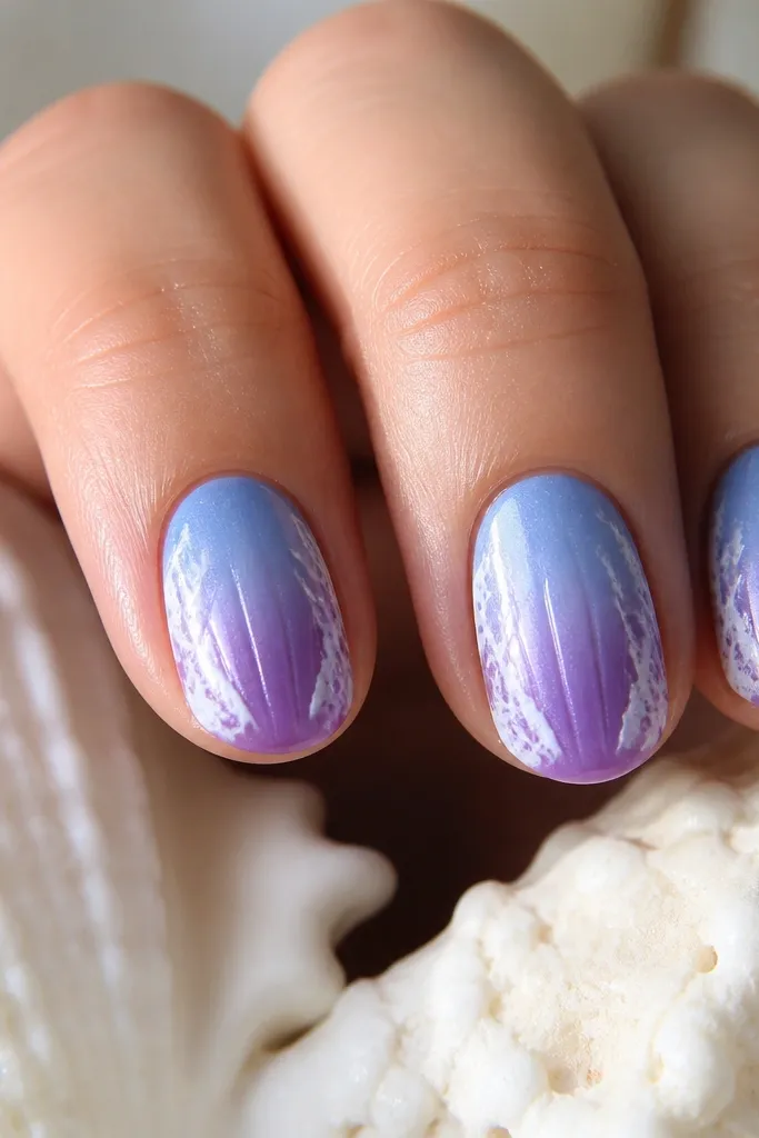 Short oval nails. Color fades from periwinkle blue near the cuticle into grape purple at the tip. Shell ridges are visible as slightly darker lavender lines running down the center. A subtle white foam flick sits along the outer ridges.
