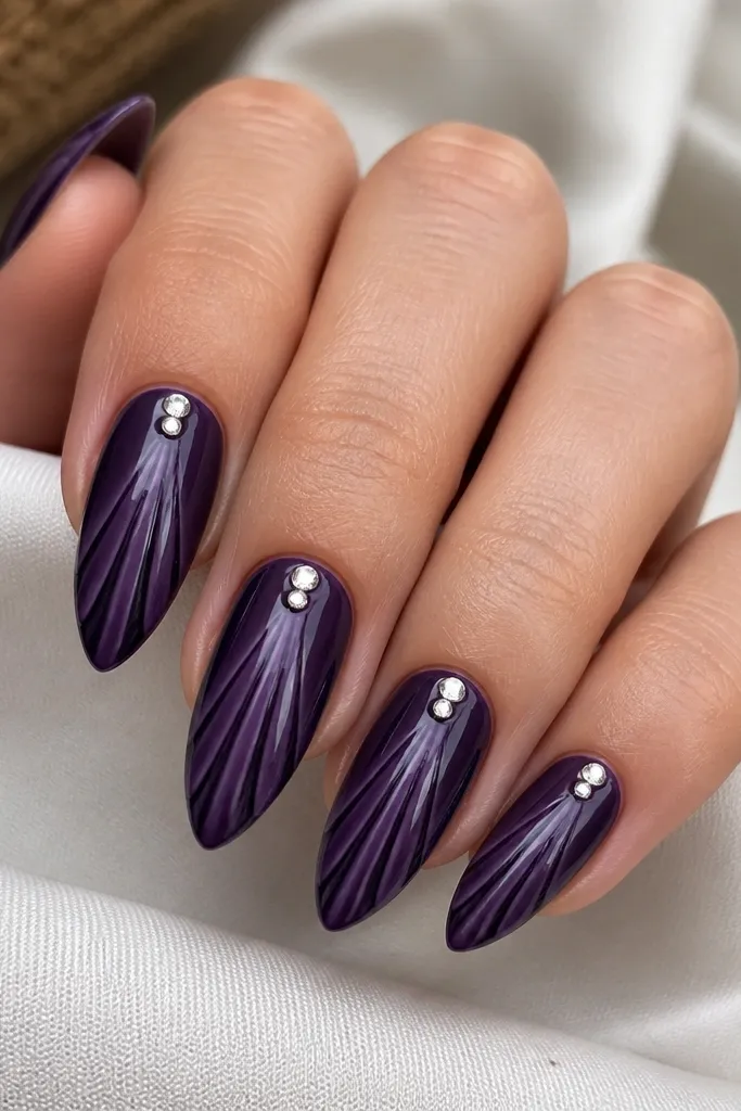 Medium stiletto nails. Base is deep grape. Shell ridges are lighter purple curved lines. Two clear, faceted mini gems sit near the cuticle on each nail, catching light. No glitter, just gems and gloss.
