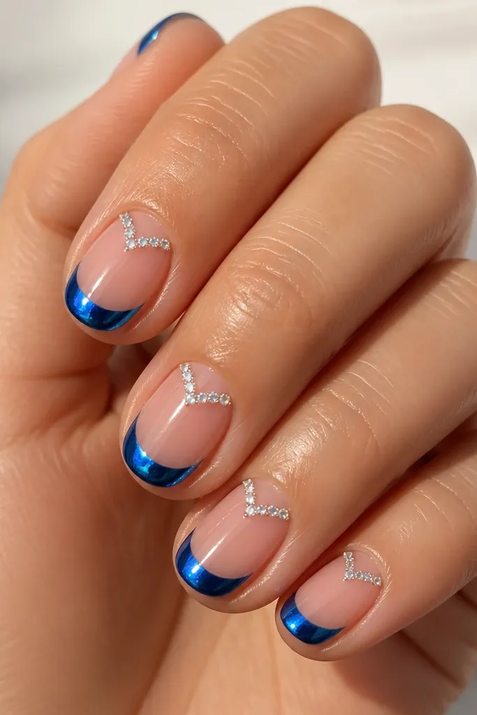 Short oval nails with bright blue chrome French tips; on each nail, tiny clear crystals form a V shape at the cuticle center; nude base, mirror shine, crystals sparkle.
