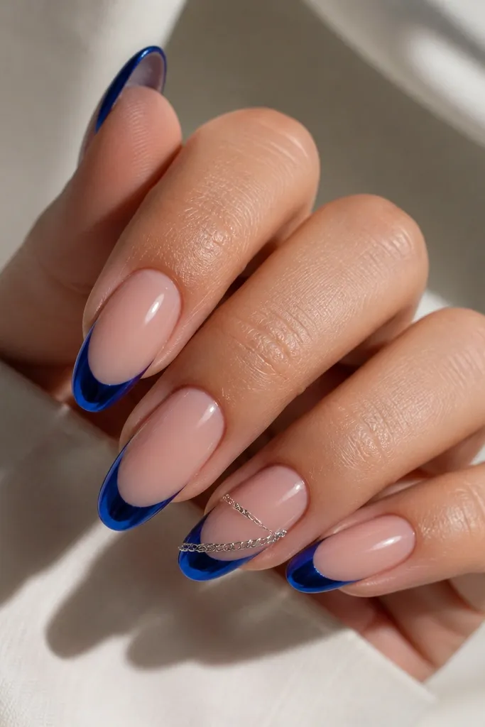 Long oval nails with royal blue chrome French tips; one accent nail has a tiny silver chain link segment running diagonally across the nude area beneath the blue line; glossy chrome, clean lines.