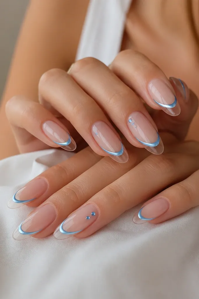 Hands with long almond nails: a thin French line in sky blue chrome sits above a see-through jelly clear tip. The chrome line looks mirror-smooth, and the clear tip shows a glossy, glassy depth. Two tiny flat blue studs sit on the outer half of the ring finger, catching light with a faceted sparkle.