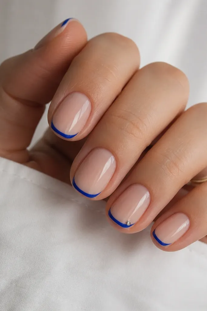 A set of short soft-squoval nails with glossy nude base. The French tips are tiny and arced, painted in bright royal blue. The smile line is thin, and the blue fades to the edges with clean tapering. One accent nail has the same micro tip with a single silver dot on the outer corner.
