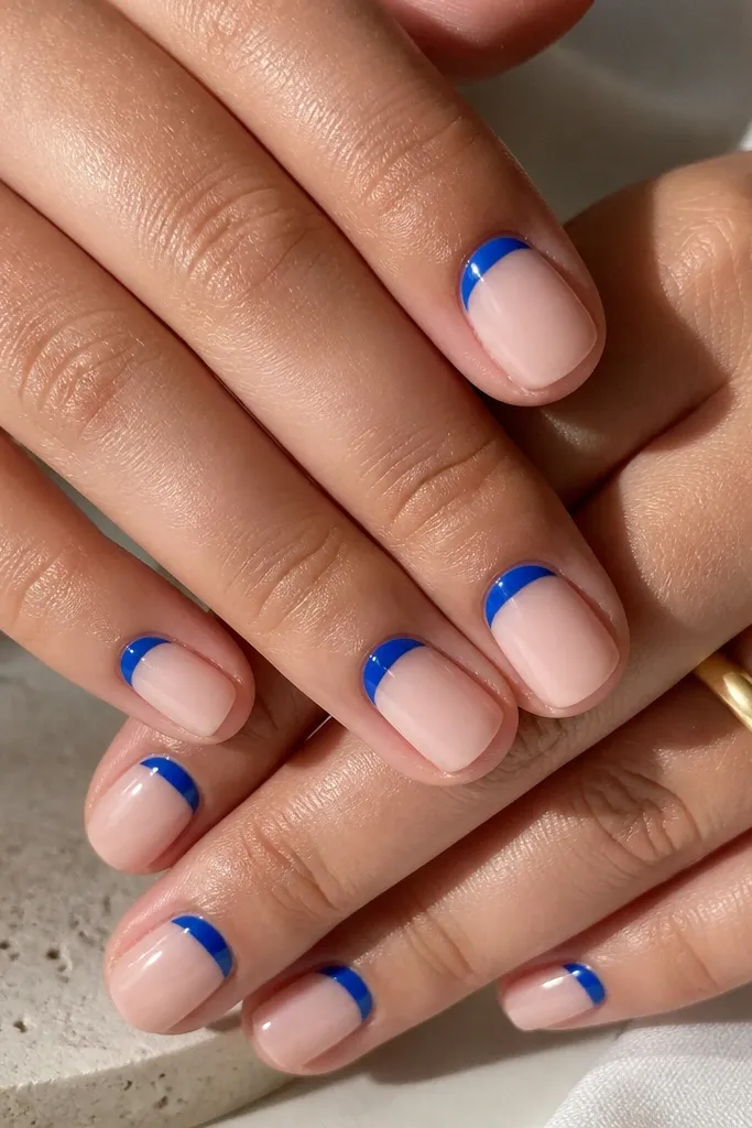 Short nails with a sheer nude base. Instead of a full French arc, there's a royal blue half-moon crescent at the cuticle area, leaving the rest nude. The crescent is crisp and glossy, with a clean curve that follows the cuticle line. The free edge is bare nude with a polished shine.