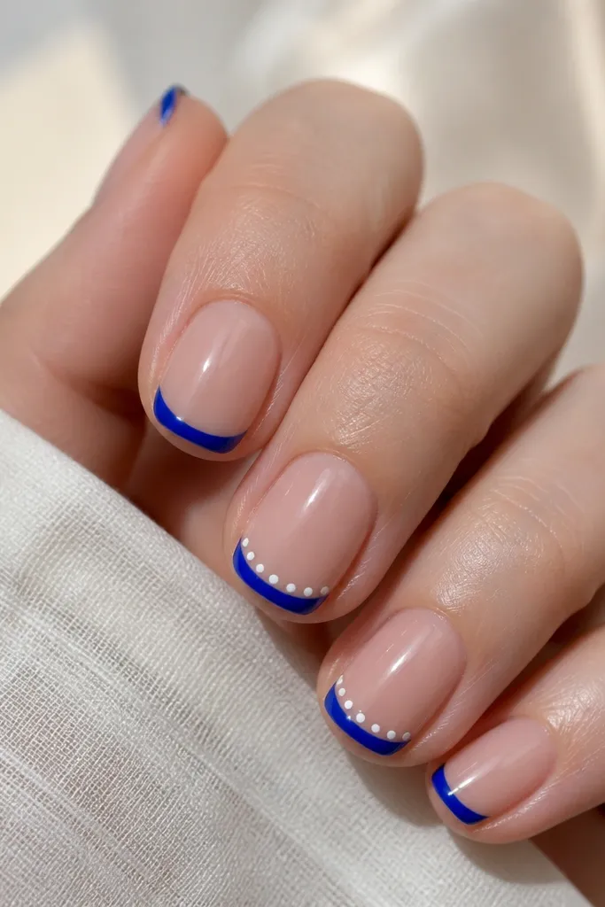 Short nails with a glossy nude base. The French tips are royal blue and narrow. A single row of tiny white dots sits just below the blue line on two accent nails, following the curve. The rest of the nails are plain royal blue French tips.