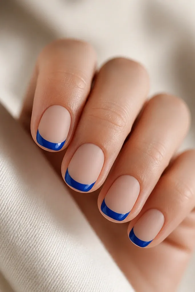 Short nails with a matte nude base. Only the French tips are glossy royal blue, with a crisp thin arc. The contrast between matte base and shiny tips is very visible under light. No extra decorations, just clean two-finish styling.
