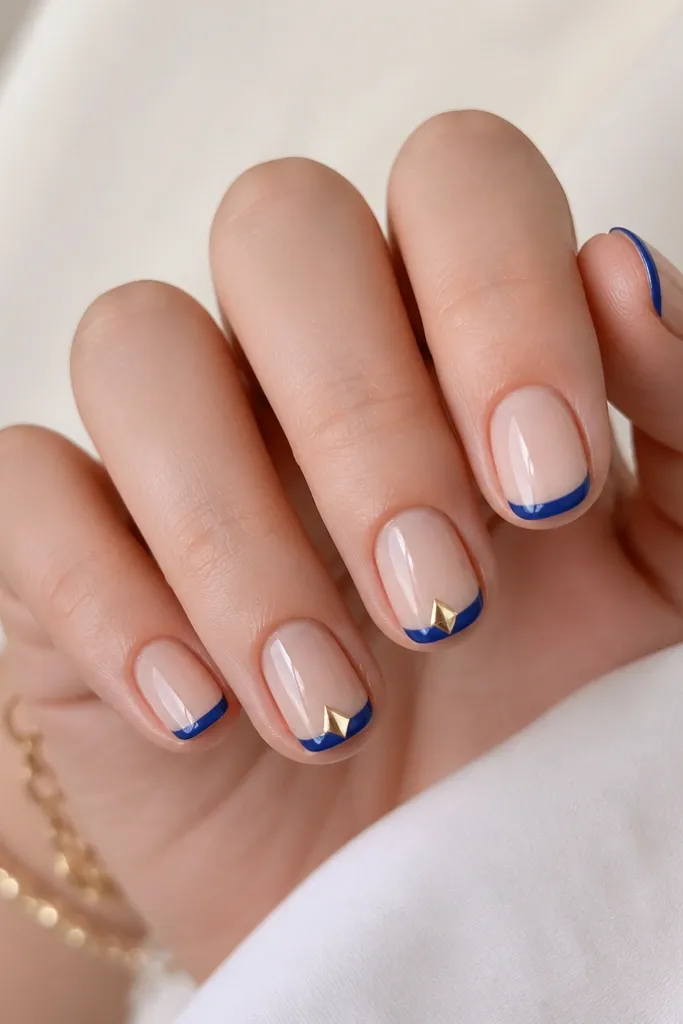 Short nails with glossy nude base. Thin royal blue French tips with a neat arc. On two nails, a tiny gold triangle sits at the center of the tip, pointing toward the free edge. The triangle is flat and metallic, with crisp edges and a mirror-like shine.