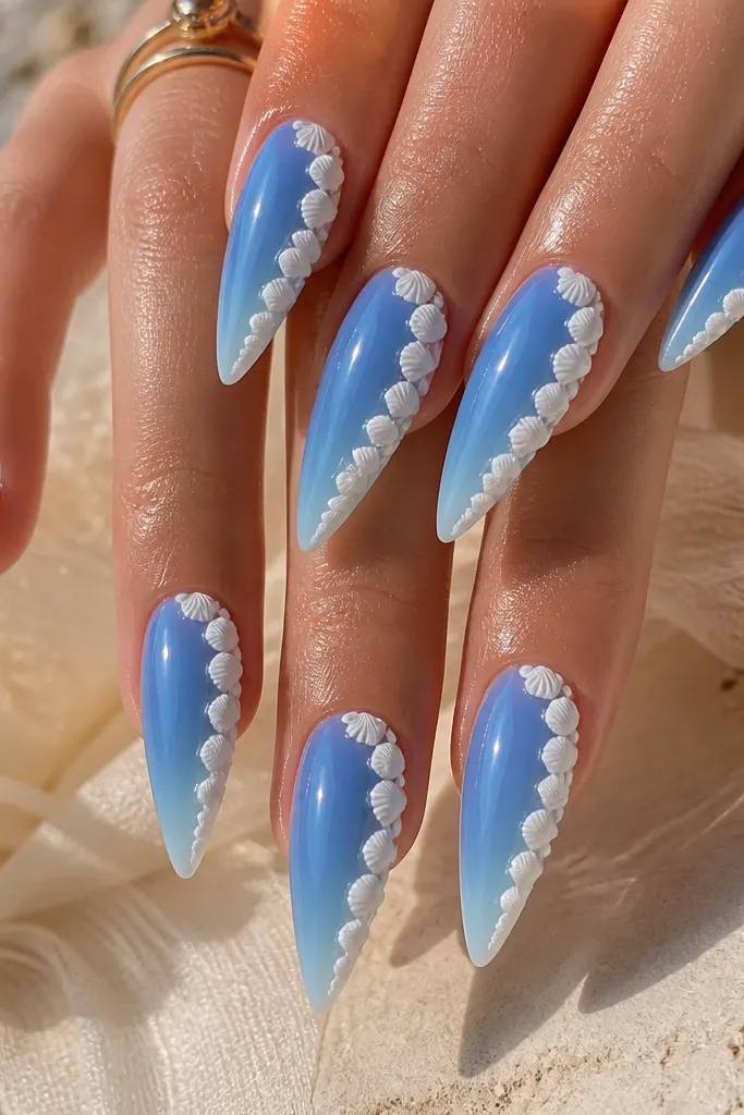 Long stiletto nails with a coastal blue base that fades slightly lighter toward the tips. On each nail, a 3D shell border runs only along the right sidewall, made of white ridged shell pieces. The left side stays mostly smooth blue. The nails are glossy and the shell border looks like a frame.