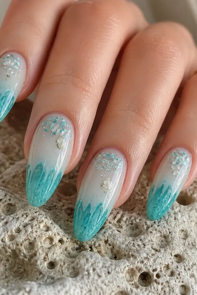 Long almond acrylic nails with a clear-to-milky sea-glass base, tiny mint and aqua confetti flakes suspended near the cuticle, and uneven turquoise drip color pooling at the tips like melted sea glass. Fine white specks and a few translucent shell flakes catch the light. Glossy top coat with crisp cuticle line.